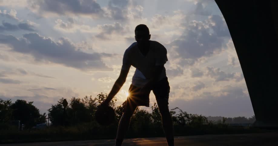 Man dribbles basketball on court partly silhouetted by sun from behind
