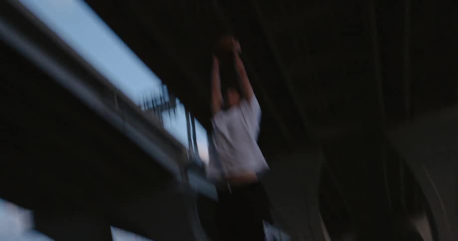 A man slam dunks a basketball in a hoop on an outdoor basketball court under a pair of bridges during the day