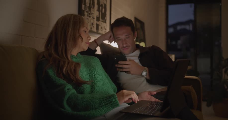 A woman uses a laptop while talking to a man using a smartphone next to her on a sofa in the living room of a house at night