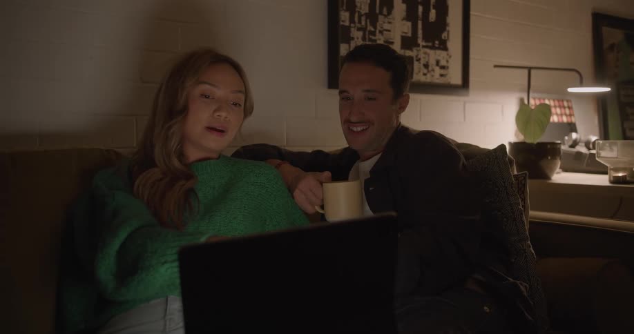 Man and woman sit on couch near desk in dark room talking while viewing laptop