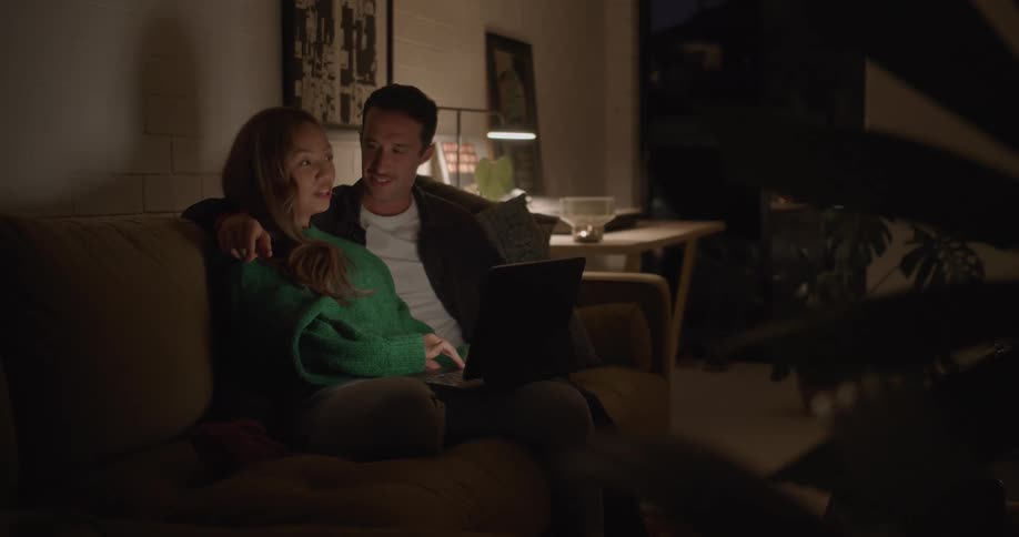 Man and woman sitting together on the couch at the living room and having a conversation while the woman is using a laptop on her lap