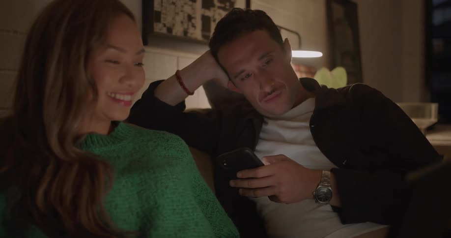 Man and woman sit on couch talking while viewing laptop and phone