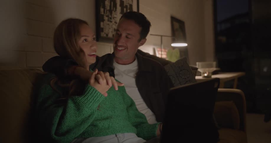 A woman using a laptop cheerfully talks with a man with his arm around her shoulders as they sit together on a sofa in the living room of a house at night