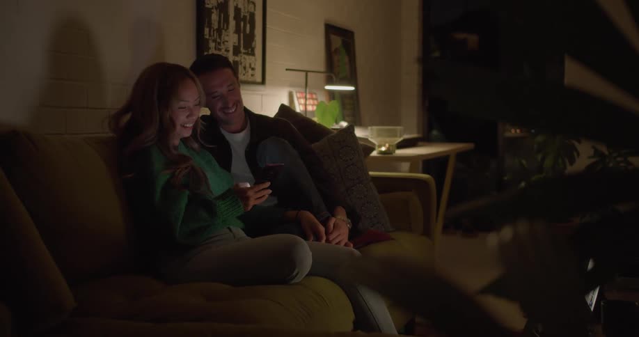 A man and woman smiling and looking at a phone while sitting close together on a living room sofa