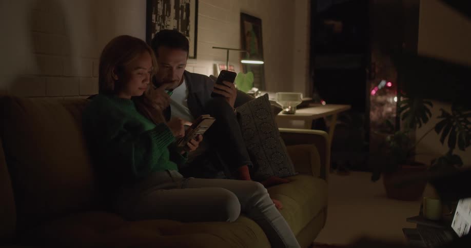 Man with mustache white and black shirt sits on brown couch next to woman with brown hair and green shirt using tablet in living room at home