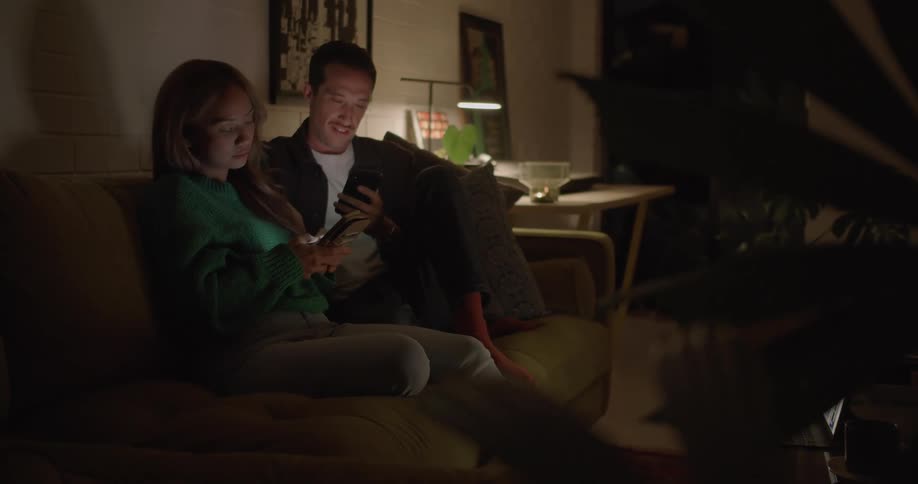 Man with moustache white and dark shirt sits next to woman with brown hair turquoise sweater and gray jeans using smartphone in living room at home