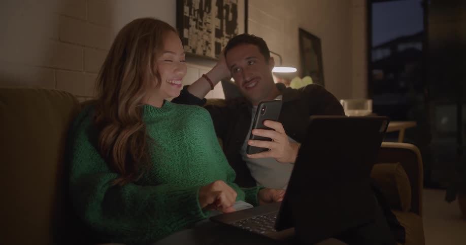 Couple sit on couch together both viewing cell phone as woman uses laptop