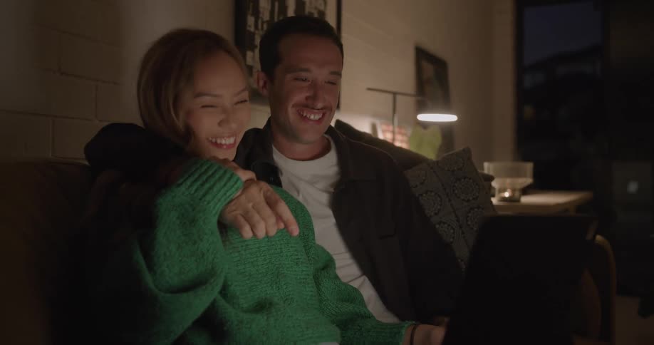 A couple laugh and cuddle on the couch while watching video on a laptop