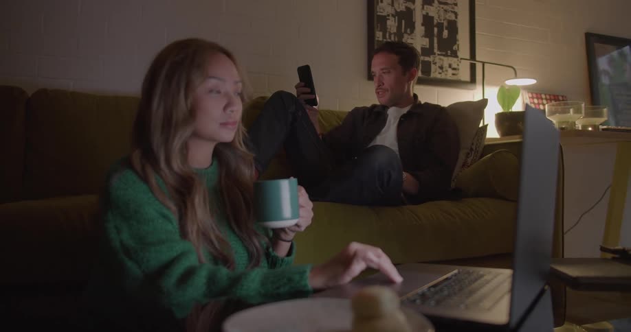 A woman using a laptop while sitting on the floor before a small table turns her head to speak to the man using his smartphone on the sofa behind her