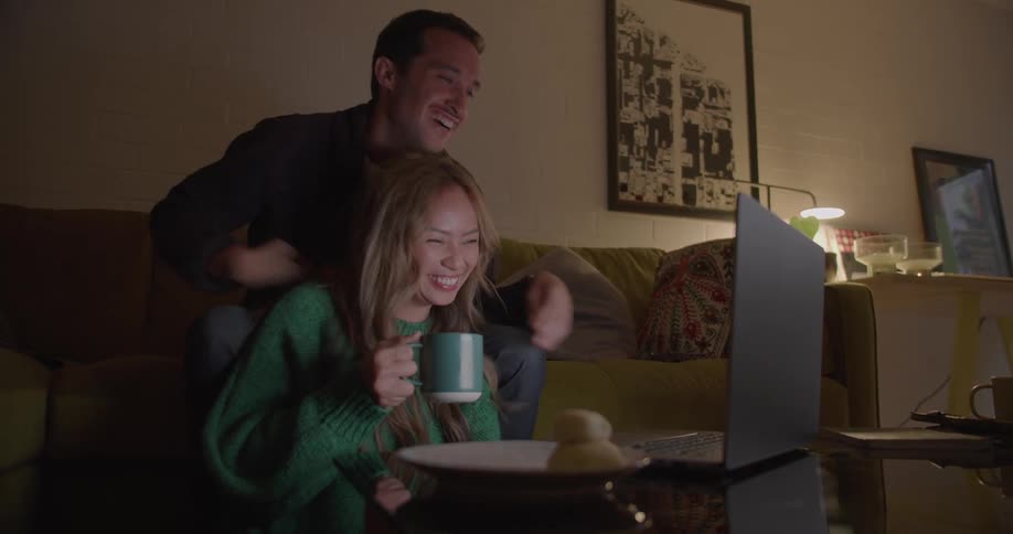 Couple sit one on the floor one on the couch and hold hands while laughing at laptop screen