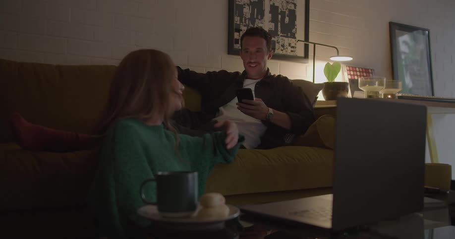 A woman watching a film on a laptop turns to encourage the man focused on his phone behind her to watch as well