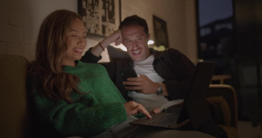 Woman points at a laptop screen as both she and her husband laugh while sitting on a sofa