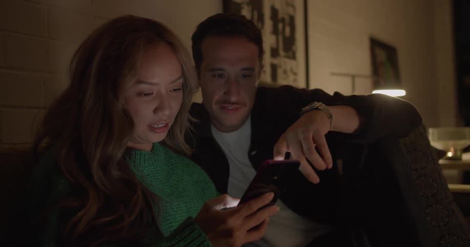 A woman holding a smartphone and the man sitting on the sofa next her look at the screen together in a dimly lit living room