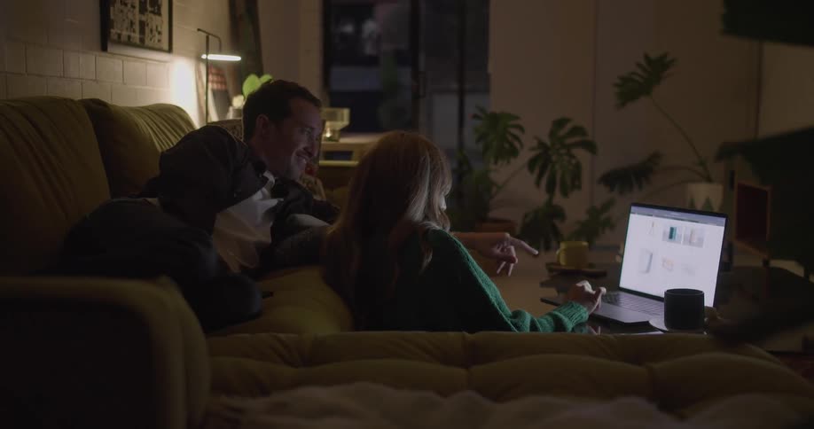 A woman sits on the floor to use a laptop on a coffee table while a man lying on the sofa behind her points at screen and speaks
