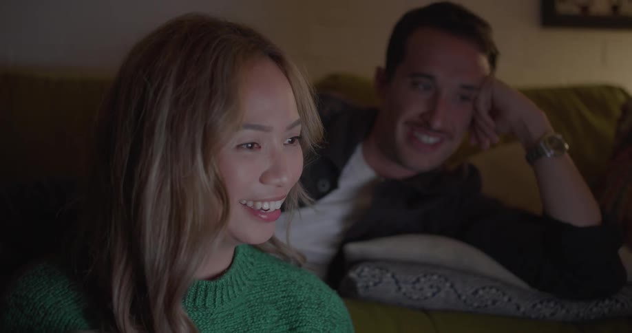 Man wearing a wristwatch relaxes on the sofa as his wife sits on the floor in front of him and they smile and laugh