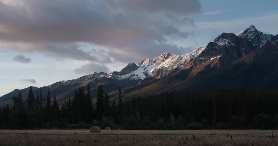 A snow capped mountain ridge looms over a field of grass holding bales of hay