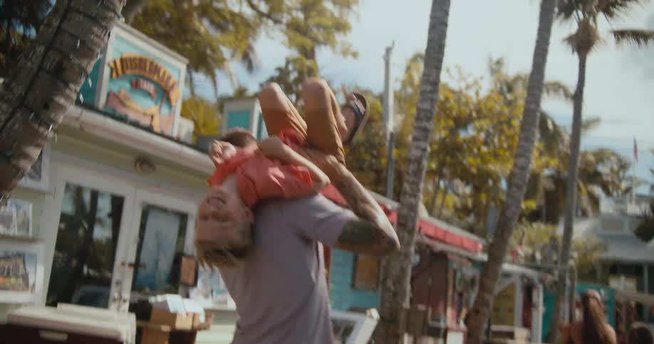 A man picks up his son and throws him over his shoulder as they walk past storefronts