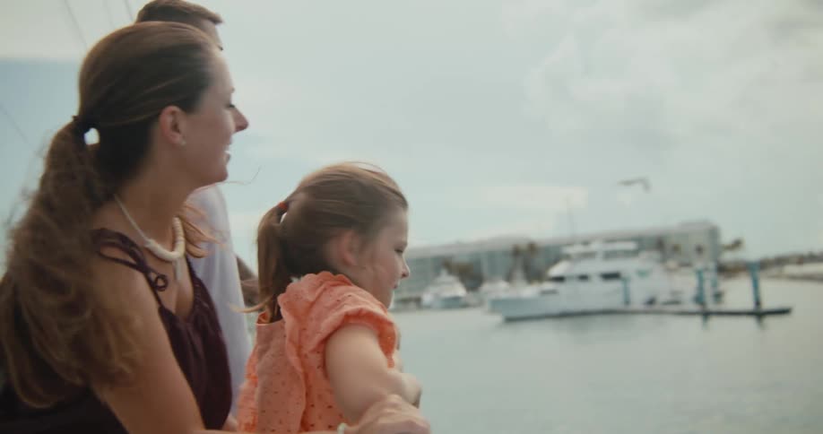 A woman and man standing on a boat point at things in the distance to a girl standing with them near the railing