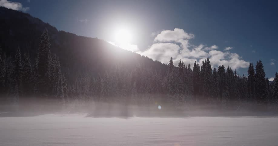 Morning fog sits over a frozen lake with sun and snowy mountains behind