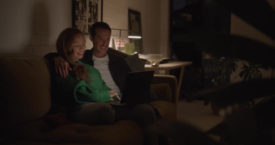 Couple sit on couch hugging and smiling while they view laptop screen and talk