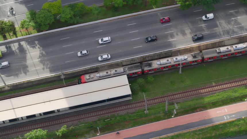 A train travels into a station near cars on highways and people riding bikes on a path around the river