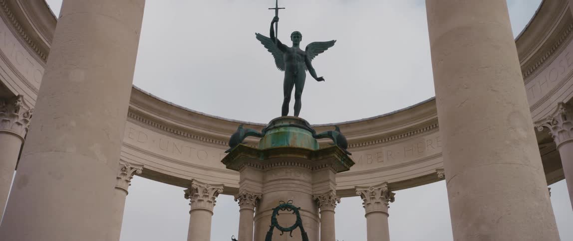 A sculpture of a man with angel wings sits on a pedestal in a open air building made of columns