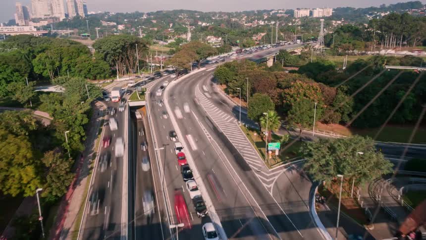 Cars and trucks travel on a city highway and under a bridge near the trees and buildings