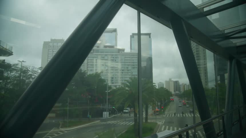 Pedestrians and vehicles move across intersection near enclosed pedestrian bridge with skyscrapers in the background