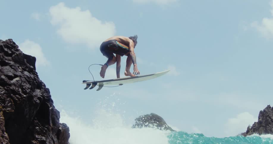 A male surfer jumps off of a rock formation