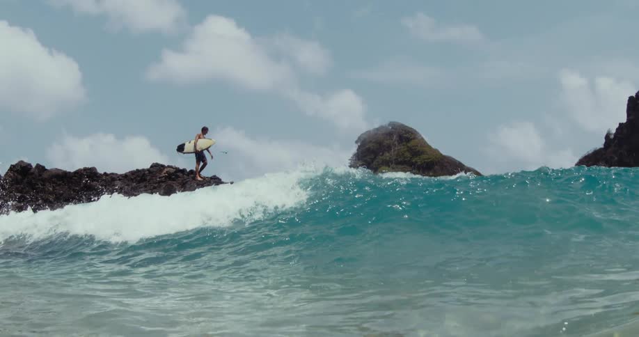 View from the water of a surfer standing on a rock holding his surfboard as a wave rolls overhead and the view is now underwater