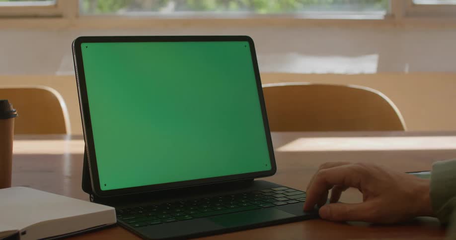 Someone is sitting at a table looking at a green tablet screen with their hand near the keyboard