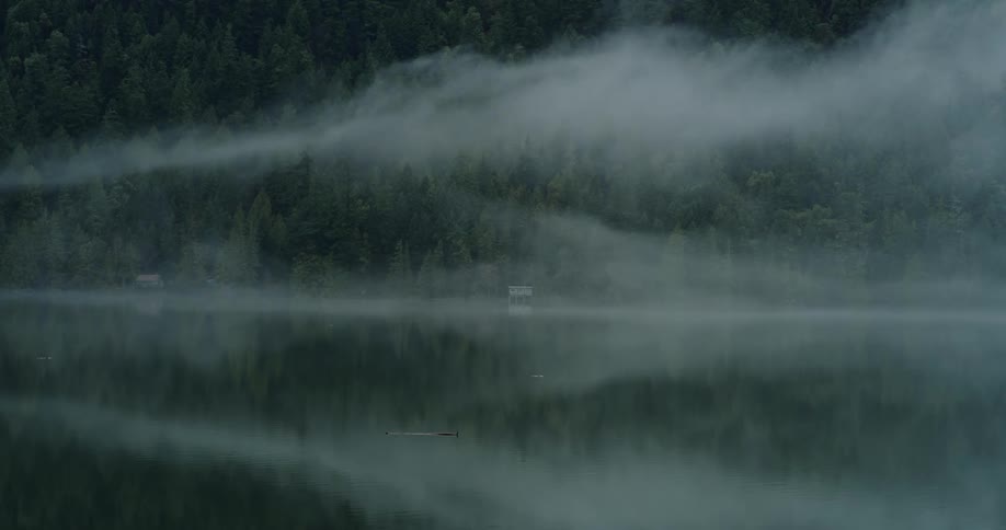 A lake at the base of a forested mountain slope shrouded in fog during the day