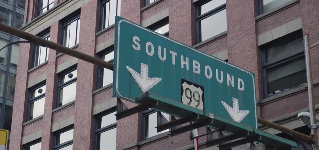 Green rectangular sign with a highway number and down arrows attached to a pole next to a tall building