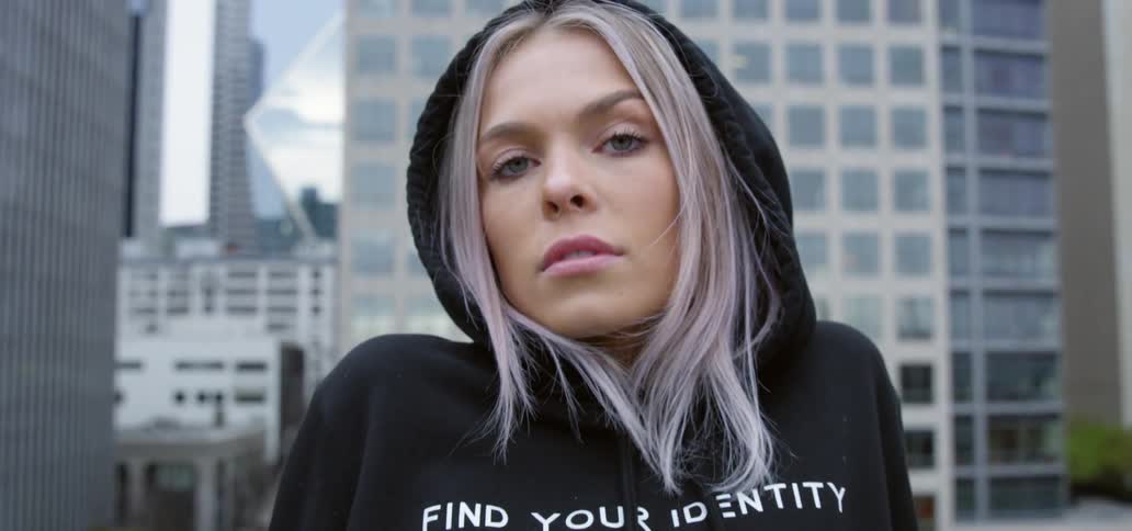 A woman wearing a hoodie poses for the camera while atop a building in an urban area