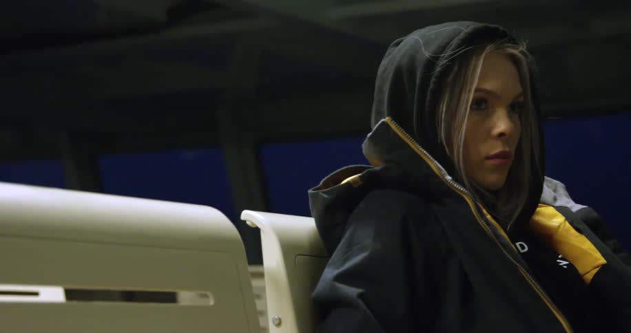 A woman wearing her hood up along with a coat sits on a plastic seat traveling at night