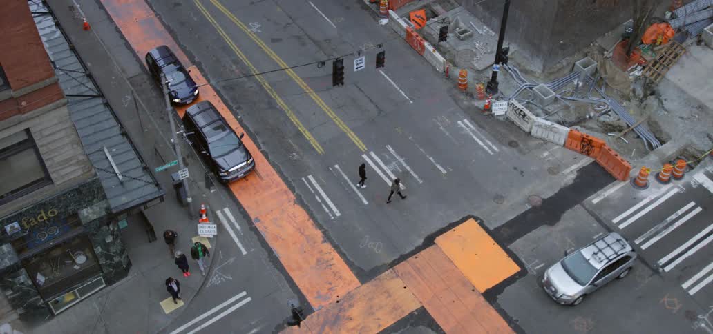 People cross crosswalk next to vehicles at intersection seen from tall building in the city