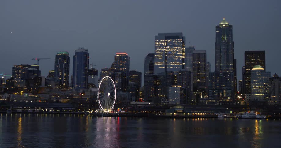 Illuminated city skyline with high rise buildings and ferris wheel seen from boat traveling on water at night