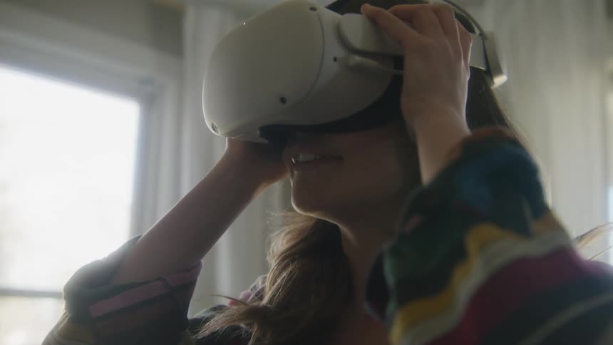 A woman smiles as she dons a white virtual reality headset while standing in a living room