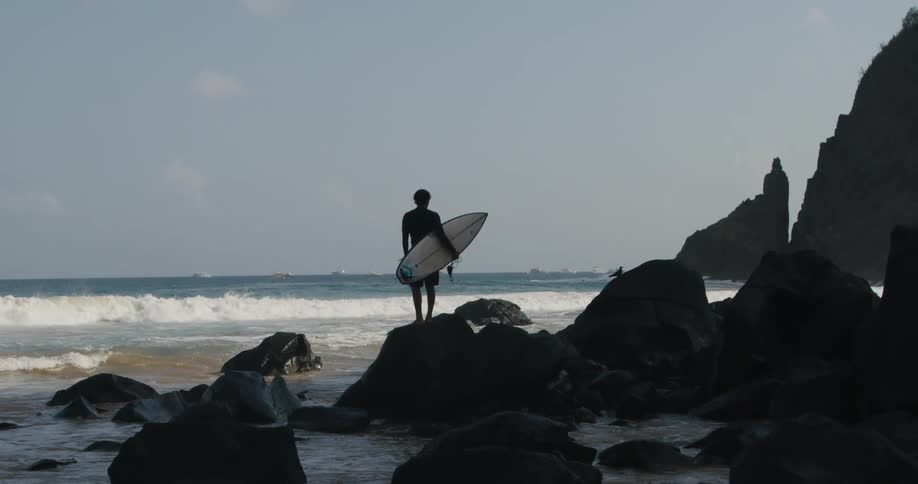 The silhouette of a man standing on a rock holding his surfboard watching the waves