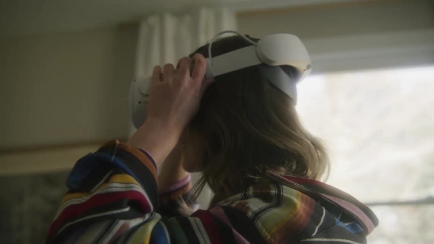 A woman places a virtual reality headset on her head while standing in a house during the day