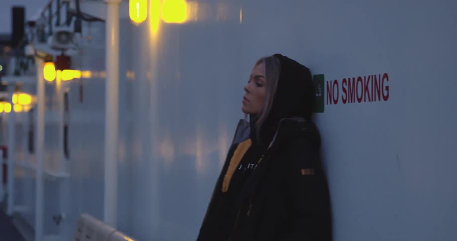 A woman leaning against a wall with a no smoking sign outside at night