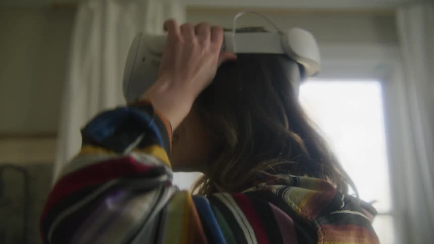 A young woman places a virtual reality headset over her face while standing indoors during the day