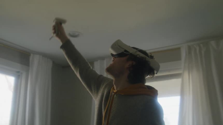 Man with gray and yellow hoodie equipped with a vr headset and motion controllers uses virtual reality at home