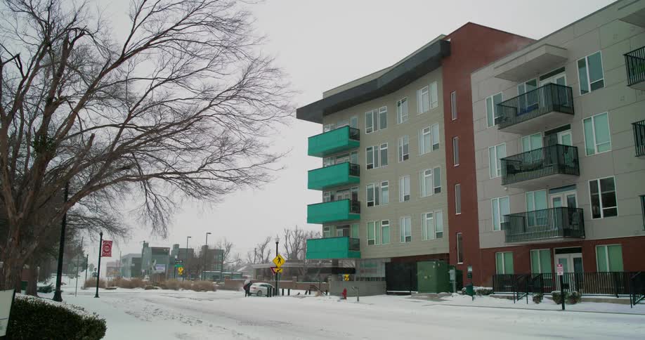 Light snow falls near an apartment building and a couple of automobiles are on the road as someone crosses the snow covered street