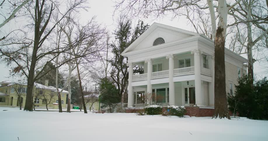 Snow falling around a white house with pillars on a winter day