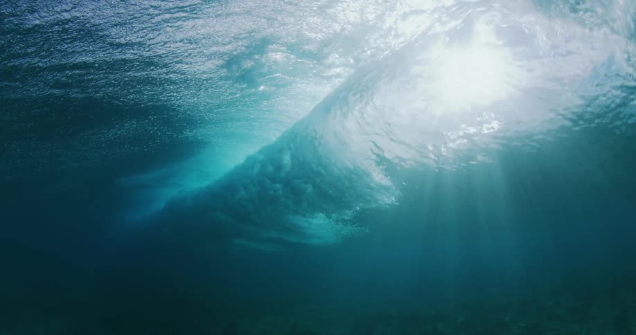 Wave rolls across surface as sun shines through surface and murky water onto seafloor