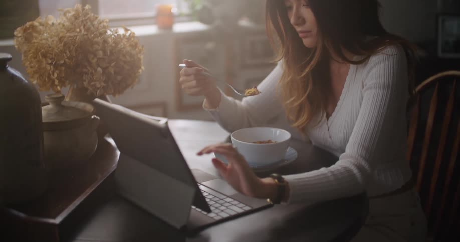 Pretty woman sitting at table eating cereal while working from laptop