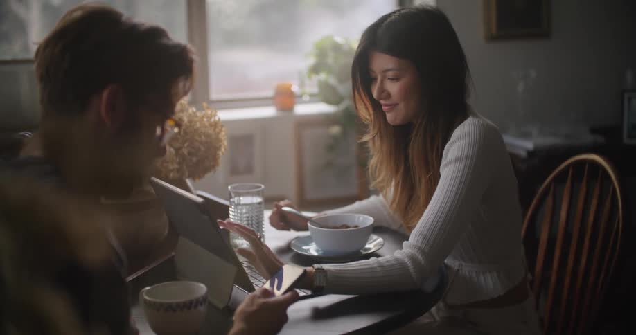 Man with glasses and beard uses smartphone next woman with brown straight hair and gray shirt working on tablet at table in the kitchen at home