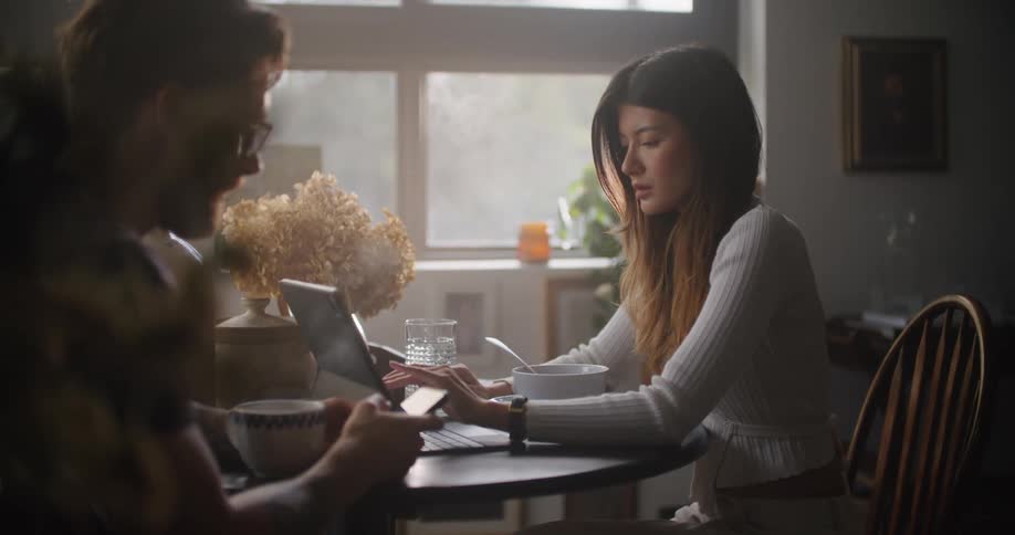 Man and woman sit at a table as the woman scrolls with her finger on a laptop screen and the man looks at his cellphone