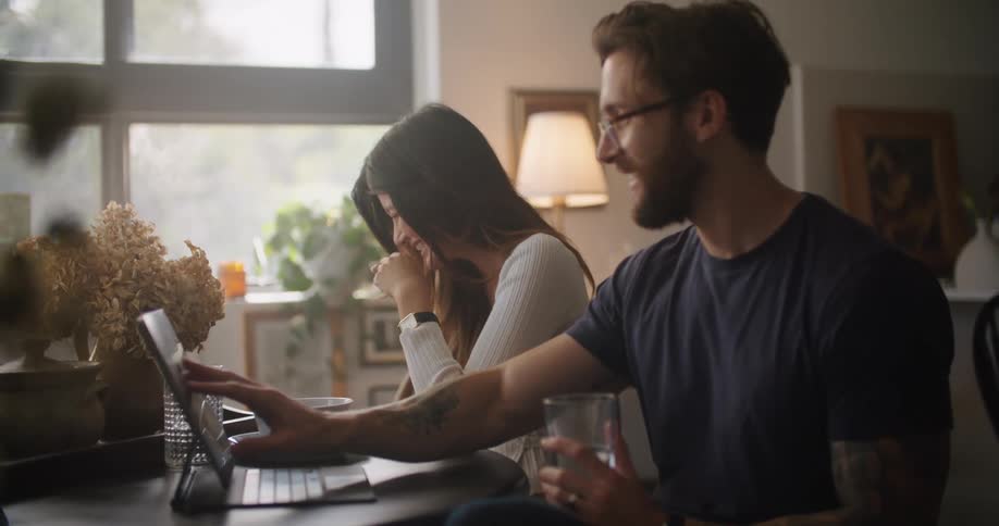 Young couple laugh as they use a tablet at the dining table in the morning